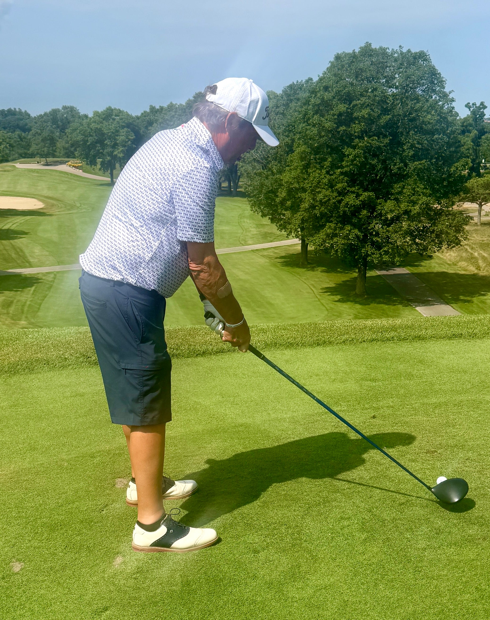 A man wearing a white patterned polo shirt, navy shorts, and a white cap is mid-swing on a golf course, hitting a golf ball with a driver. The background shows a lush green golf course with trees under a blue sky.