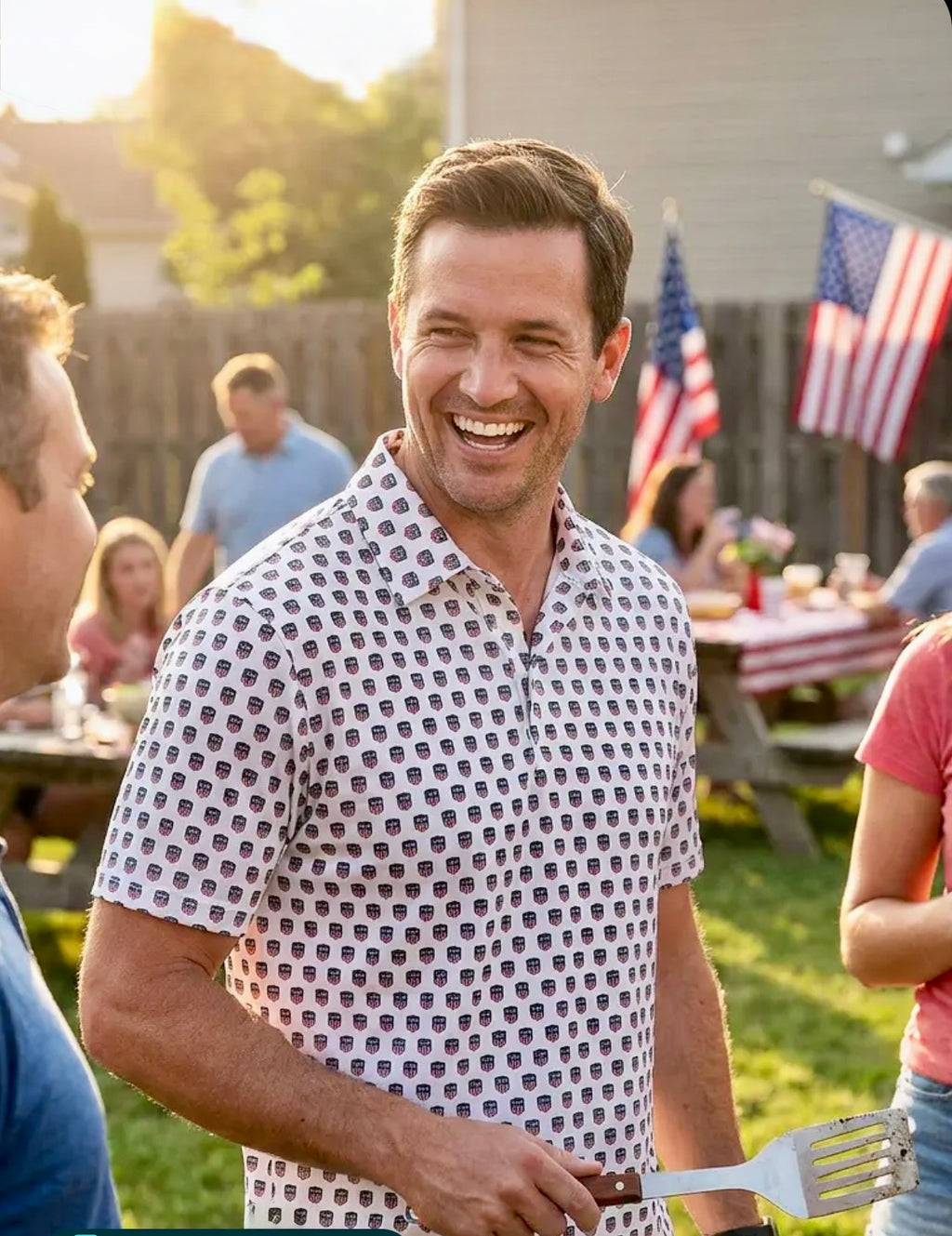 Man in a USA250 Freedom Shield shirt holding a spatula at a casual outdoor gathering with American flags in the background.