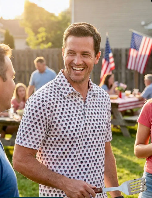 Man in a USA250 Freedom Shield shirt holding a spatula at a casual outdoor gathering with American flags in the background.